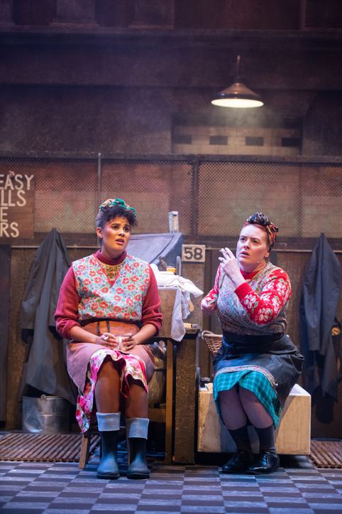This is a promotional image from a theatrical production of The Steamie by Dundee Rep. Two actresses sit side by side in floral smocks, both smoking cigarettes.