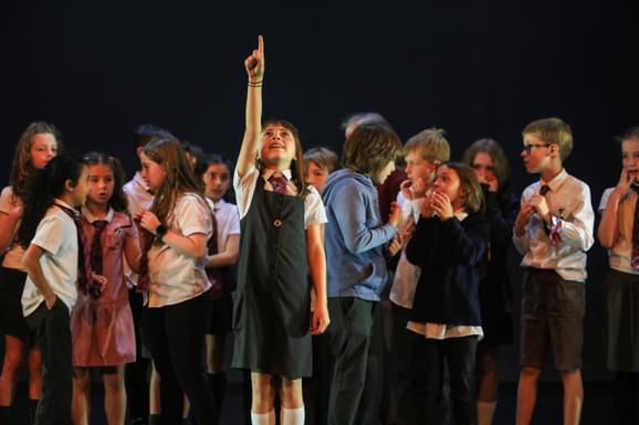 A girl stands in school uniform pointing towards the sky smiling with a group of children behind her chatting amongst themselves