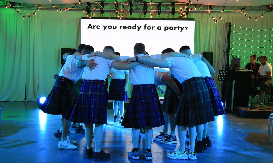 An image of a group of men wearing kilts huddled in a cirlce on a dancefloor from a performance of Beside Me
