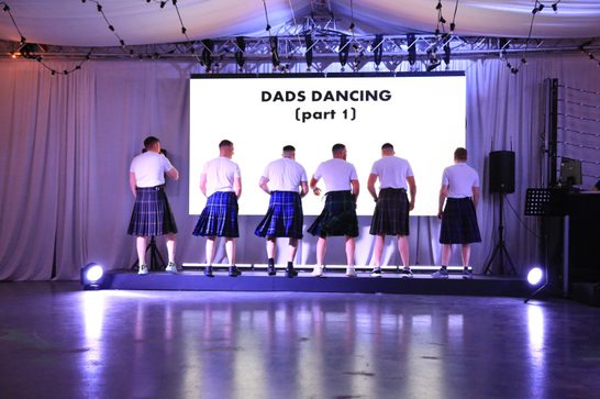A group of men face away from camera wearing kilts and white tshirts from a performance of Beside Me