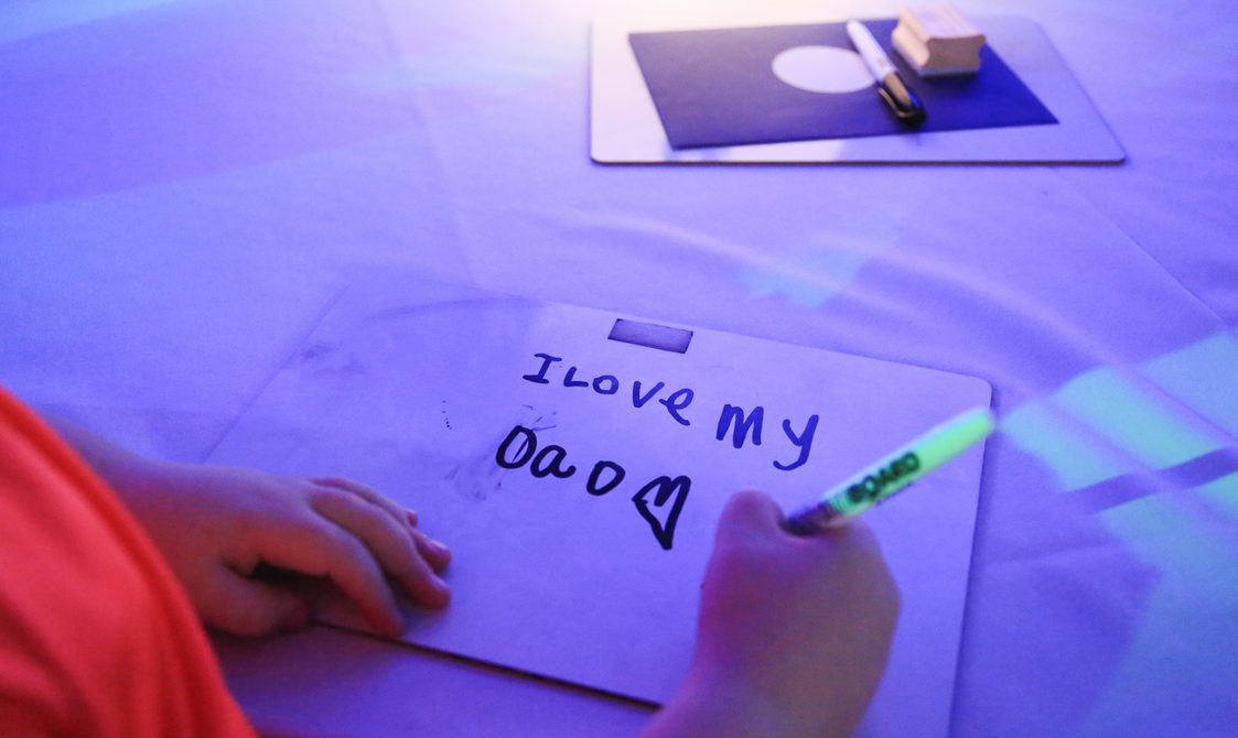 A child's hands with a pen write, I love my Dad on a whiteboard, from a performance of Beside Me