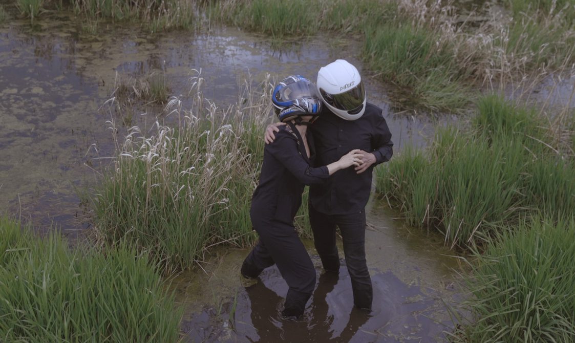 Two people stand in a swamp wearing all black and bike helmets holding onto one another