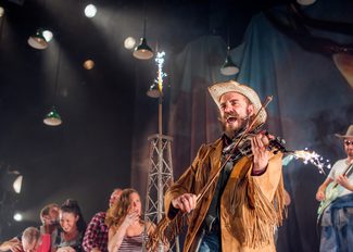 A man in a cowboy outfit is singing and playing the fiddle on stage with other performers in background.