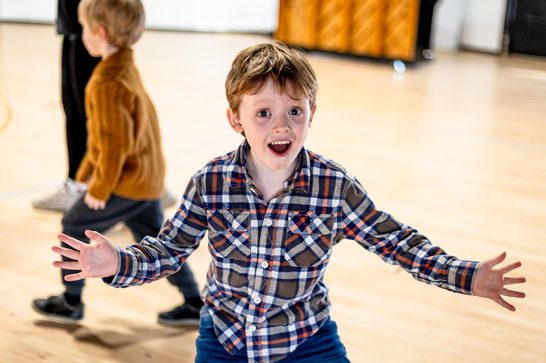 A young boy with ginger short hair wearing a checked shirt, arms outstretched and smiling