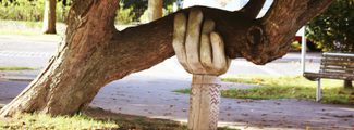 A tree trunk supported by a stone sculpture of a human hand, positioned in a park setting with a path and bench visible in the background.