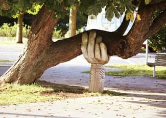 A tree trunk supported by a stone sculpture of a human hand, positioned in a park setting with a path and bench visible in the background.