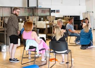 two groups of people sit in circles of chairs in conversation in a brightly lit rehearsal space