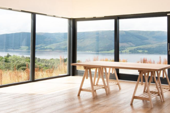 A table in a brightly lit room sits on a wooden floor in front of large glass windows overlooking a loch and rolling hills