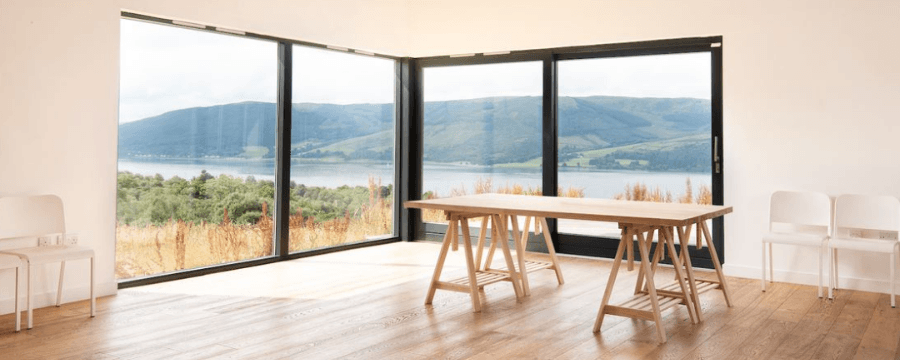 A table in a brightly lit room sits on a wooden floor in front of large glass windows overlooking a loch and rolling hills
