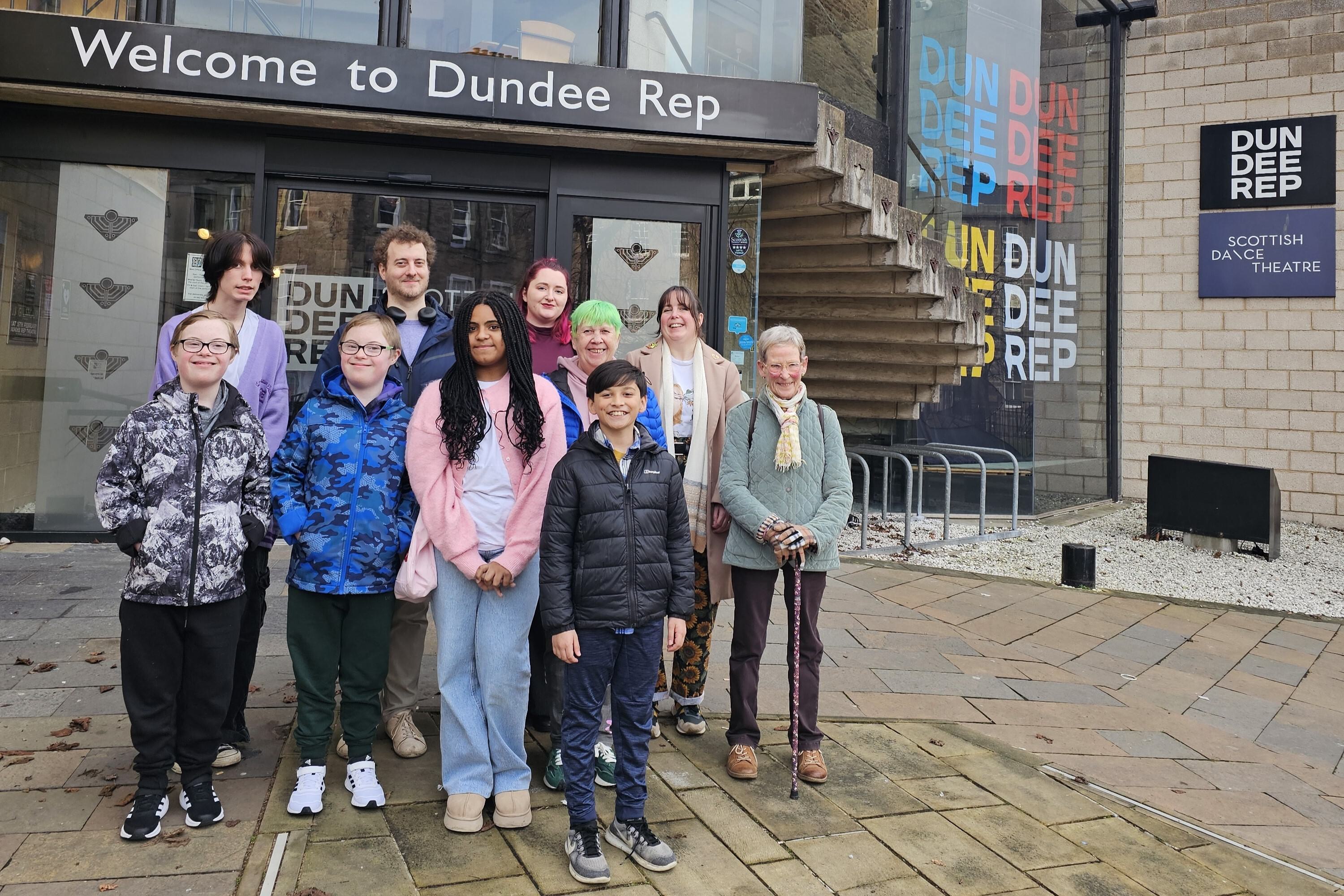 A group of ten people of varying ages and appearances pose together outside Dundee Rep Theatre, with a welcoming sign above the entrance and modern architecture in the background.