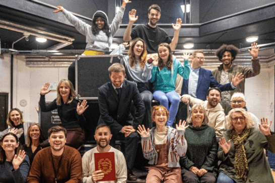 The cast of A Christmas Carol smile arranged on a set of stacked flight cases