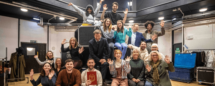The cast of A Christmas Carol smile arranged on a set of stacked flight cases