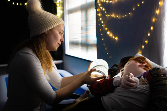 A woman in a winter hat reading to a child lying down, with soft lighting and fairy lights in the background