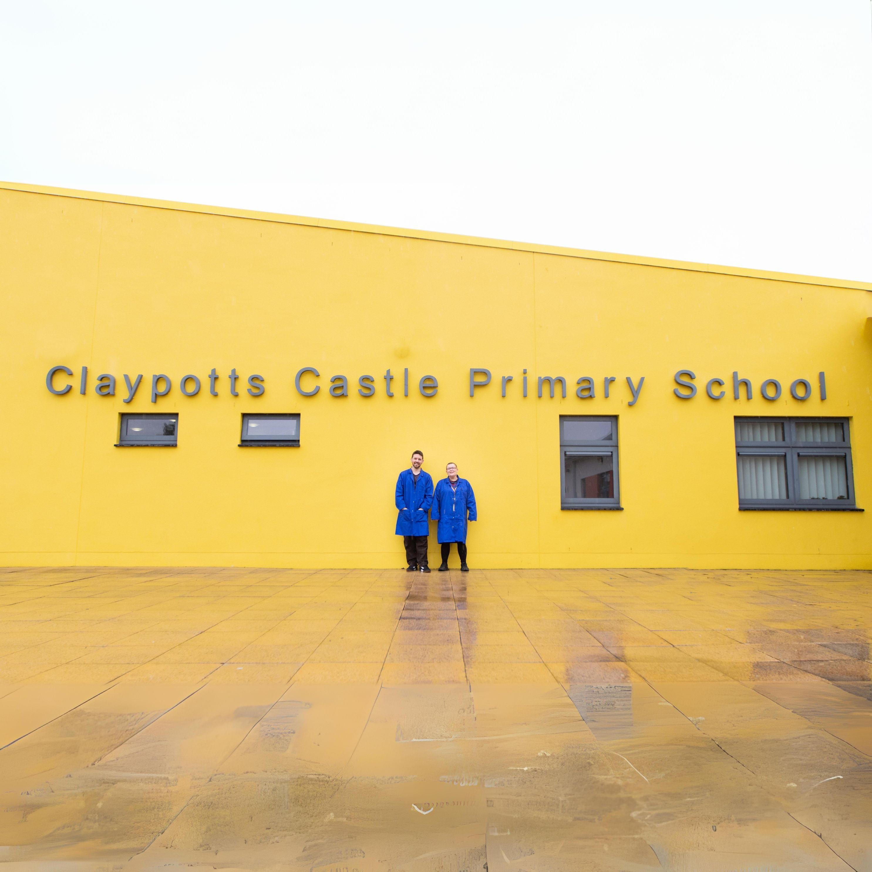 Two people in blue coats standing in front of a yellow building labeled "Claypotts Castle Primary School
