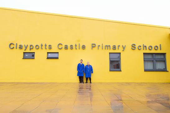 Two people in blue coats standing in front of a yellow building labeled "Claypotts Castle Primary School