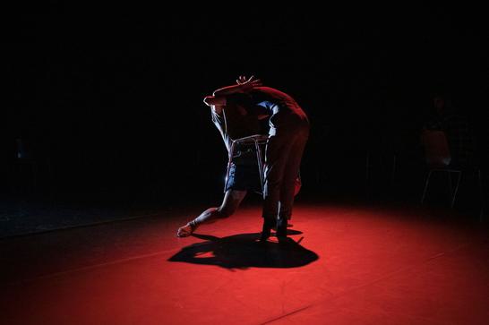 two dancers lit in red with a black background are mid-performance, closely intertwined together
