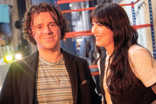 A man stands lit in orange with brown hair wearing a black jacket and striped tshirt, as a woman with long dark hair stands next to him looking at him smiling