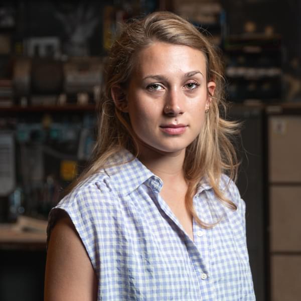 Olivia Arizemendi, a woman with shoulder length blonde hair wearing a sleeveless blue and white checked shirt looks seriously at camera