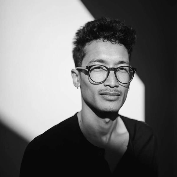 Tao Anas le Thanh, a man with brown curly hair wearing glasses and a black top looks seriously into camera