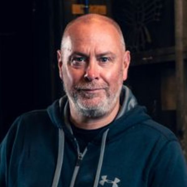 Barrie Hunter, a white bald man with a grey beard wearing a blue hoodie smiles sternly into camera