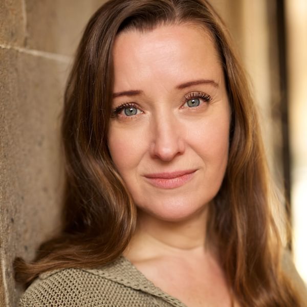 Emily Winter, a woman with long brown hair smiles into camera, leaning on a wall