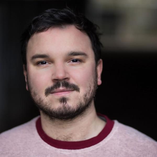 Headshot of Finn den Hertog, a man with dark hair, beard and moustache wearing a light pink tshirt with a dark red collar
