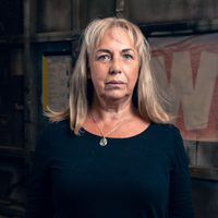 Irene Macdougall, a woman with long blonde/white hair wearing a blue top and pendant necklace stares seriously at the camera