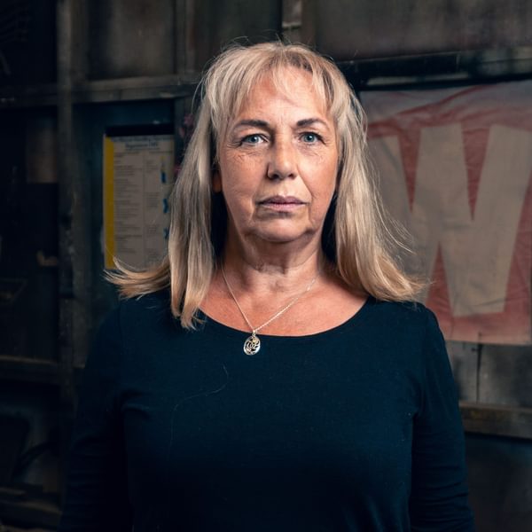 Irene Macdougall, a woman with long blonde/white hair wearing a blue top and pendant necklace stares seriously at the camera