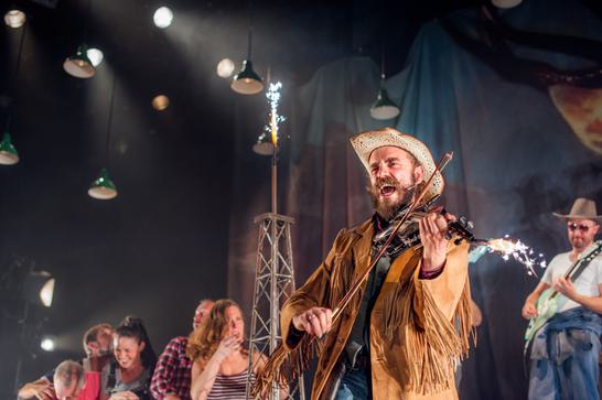 A man in a cowboy outfit is singing and playing the fiddle on stage with other performers in background.